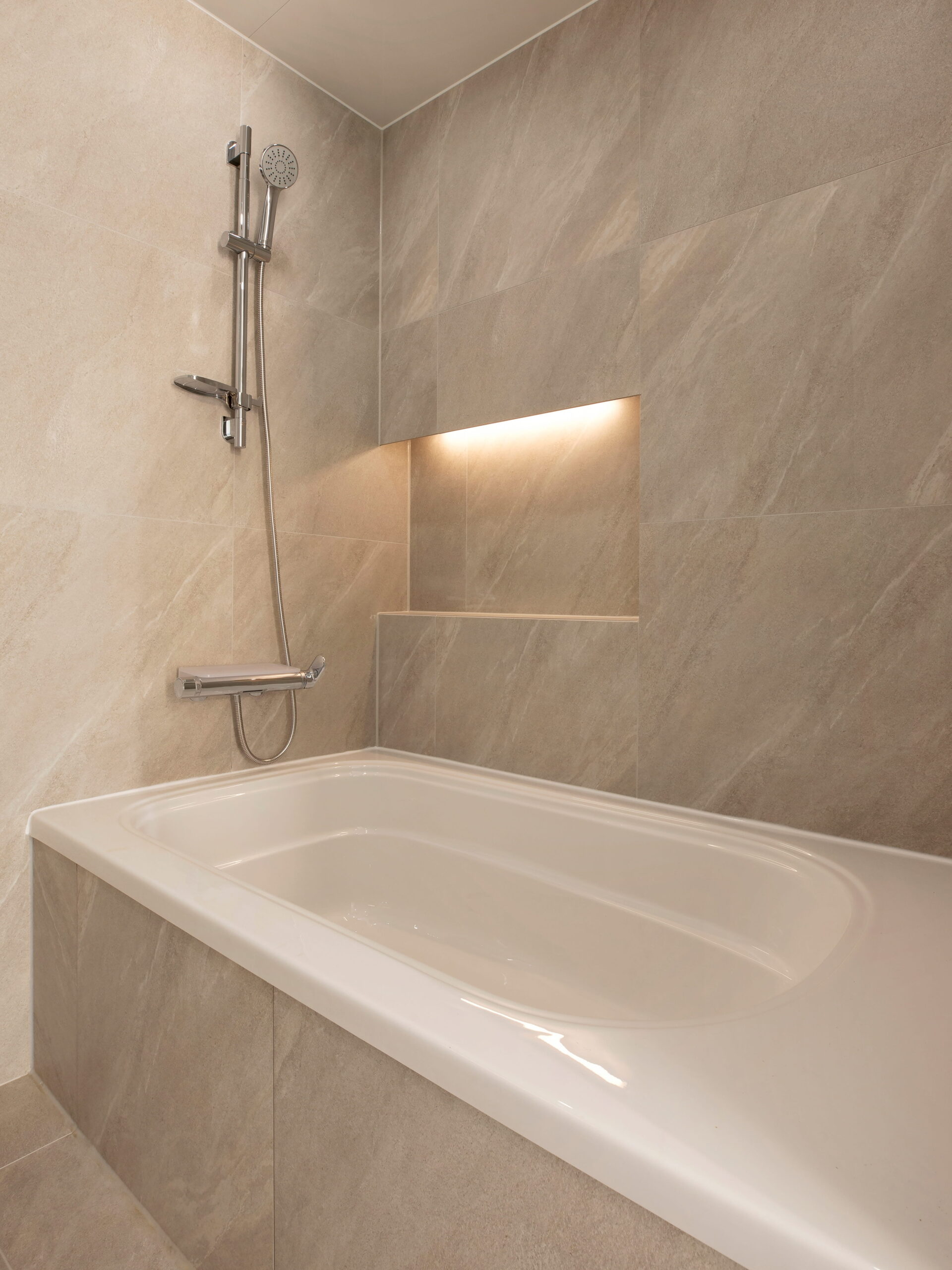 Modern bathroom with beige marble tiles, a built-in white bathtub, a sleek chrome showerhead, and a softly lit alcove, creating a serene ambiance.
